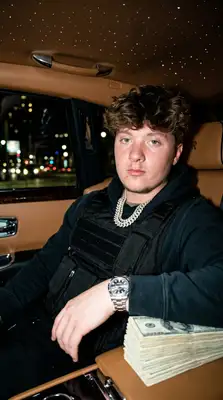 Take the uploaded photo of the person and generate a photorealistic 9х16 image of this person (please make sure to keep facial similarity) in one of the following randomly selected settings:   Leaning against a bright Lamborghini or Rolls-Royce, wearing a tactical vest or puffer jacket, showing off a luxury watch or chain.  Inside a private jet with stacks of cash on their lap, dressed in designer streetwear (like BAPE, Louis Vuitton, or Nike Tech).   Posing in front of a private jet at night, holding fanned-out stacks of hundred-dollar bills, with a confident or nonchalant expression.   Sitting in a luxury car with a starlight ceiling, wearing iced-out chains and sunglasses, mid-conversation or staring coolly ahead.   Standing against an urban graffiti backdrop, throwing up hand gestures while wearing a designer scarf or hoodie, heavy jewelry, and rings.   The final image should have a high-contrast, bold, Instagram-worthy aesthetic. Use realistic lighting and textures. Make sure the background, outfit, and pose reflect confidence, wealth, and a street-smart vibe. Rotate between settings randomly each time this is run.